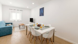 Un espace de repas moderne avec une table blanche dressée pour quatre personnes, entourée de chaises blanches. Un canapé bleu est placé à gauche, et une petite table en bois se trouve près de la fenêtre.
