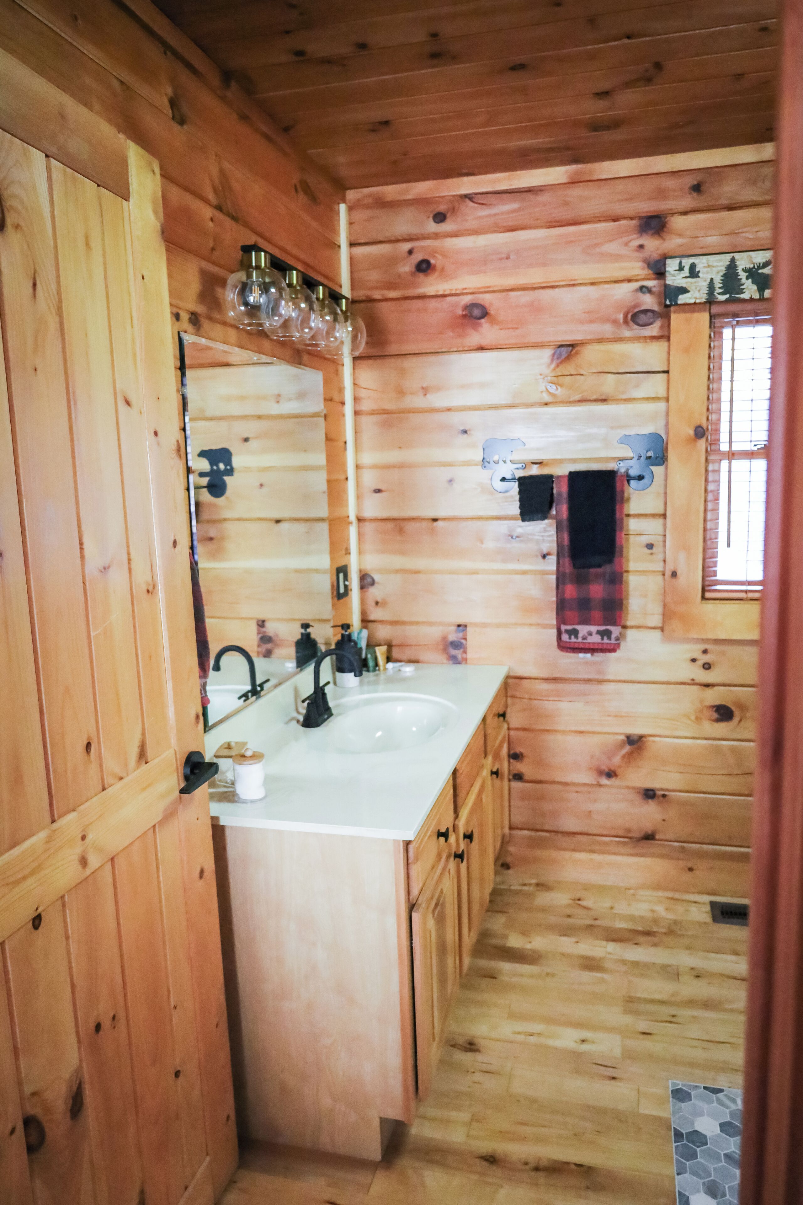 A wooden cabin bathroom with a white sink, wooden cabinets, a mirror, and a window with a plaid towel hanging on the wall.