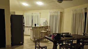 Een ruime woonkamer met een eettafel, stoelen, een bank, een keuken met een koelkast en een plafondventilator.