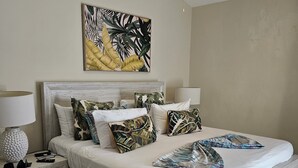 Een moderne slaapkamer met een groot bed, een plafondventilator, een nachtkastje met lamp en een schilderij met een tropisch thema aan de muur.