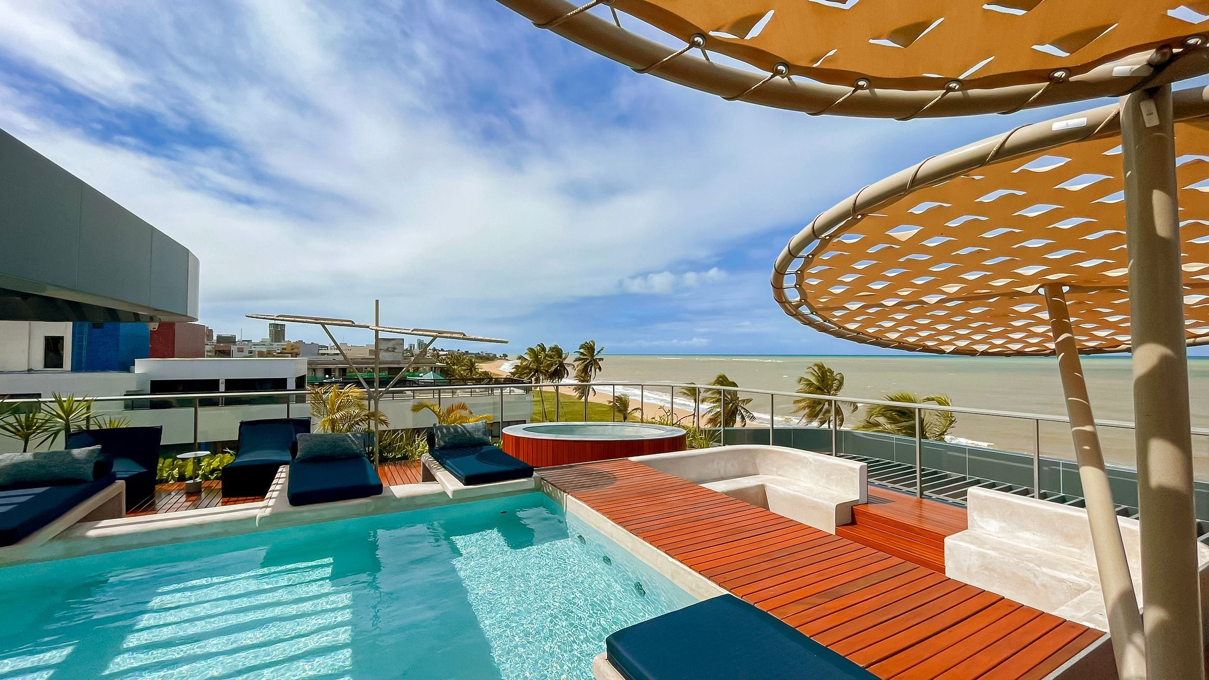A pool area with lounge chairs and a hot tub, overlooking a beach with palm trees and a clear sky.