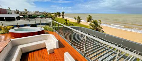 Eine Dachterrasse mit Whirlpool, Holzterrasse und Glasgeländer mit Blick auf einen Strand.
