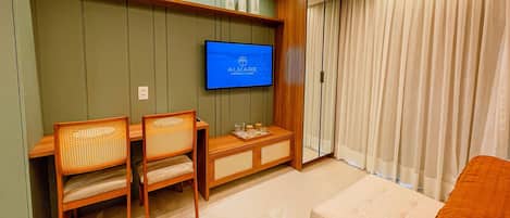 Hotelkamer met een aan de muur bevestigde televisie, een houten bureau met stoelen en een groot raam met gordijnen.