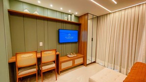 A hotel room with a television mounted on the wall, a wooden desk with chairs, and a large window with curtains. - Aquamaris Luxo - by Almare (João Pessoa)