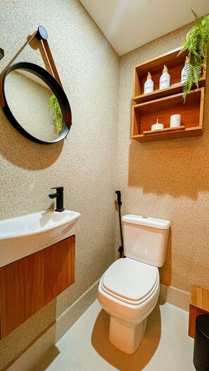 Une salle de bain avec un lavabo blanc, des toilettes, une étagère en bois où sont rangés les articles de toilette et un miroir.