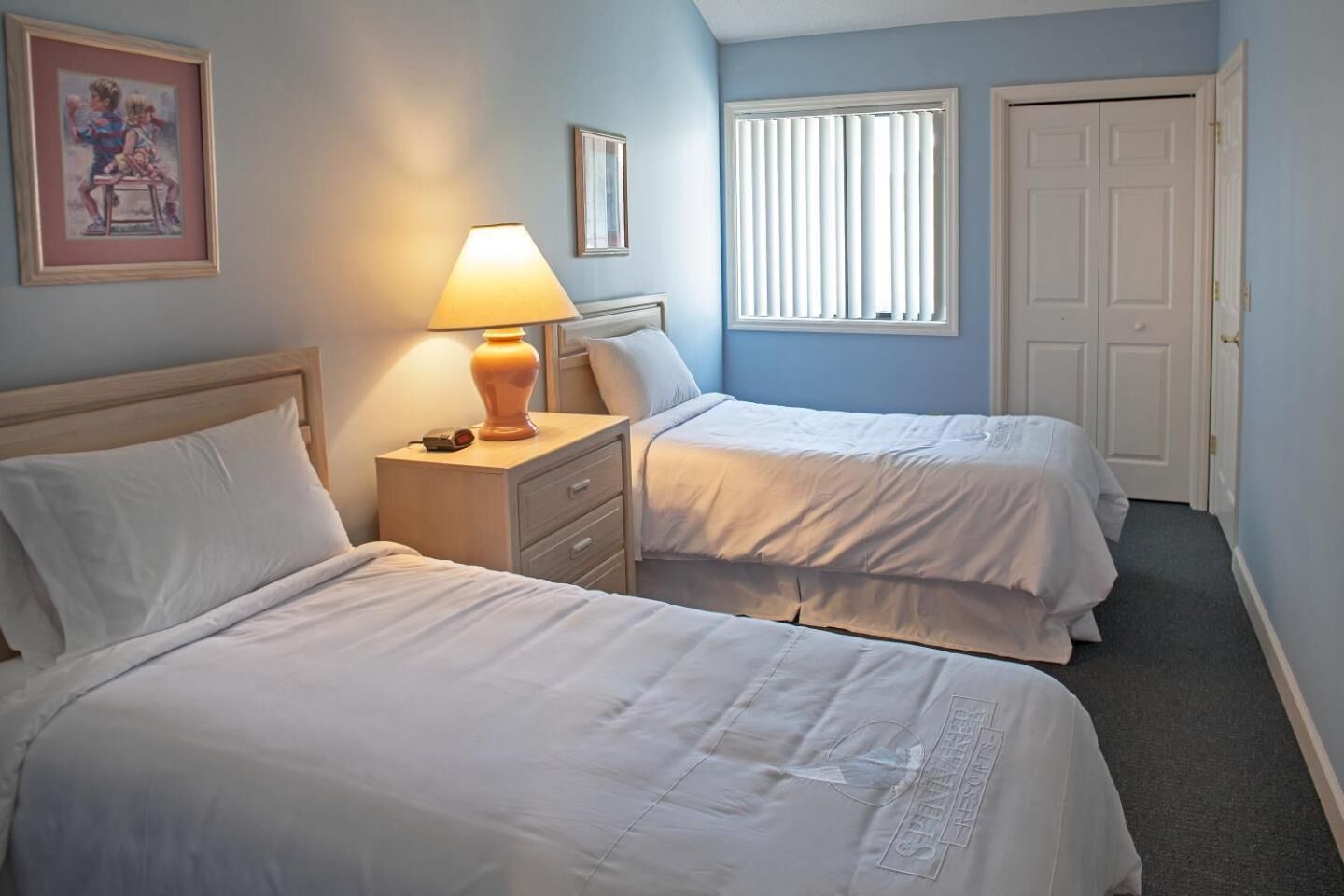 Ein Hotelzimmer mit zwei Betten, einem Nachttisch mit Lampe, einem gerahmten Bild an der Wand und einem Fenster mit Jalousien.