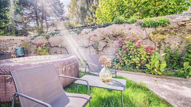 An outdoor seating area with a wicker sofa and metal chairs, a glass table with a vase, and a stone wall with greenery and flowers.