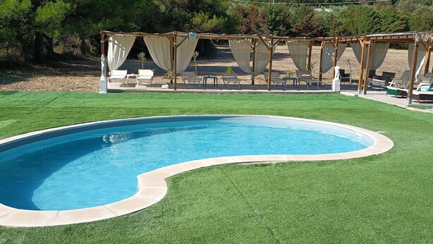 A swimming pool with a unique shape, surrounded by artificial grass and a pergola with white curtains. - Bed and breakfast il rosmarino  (Bosa)