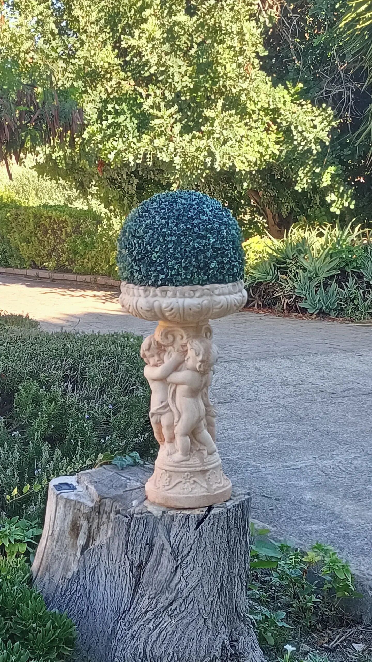 A garden statue with a topiary on a pedestal, set in a landscaped area with various plants and a pathway.