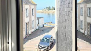 A view through a window of a residential street with houses, a parked car, and a body of water in the background. - Appartement idéal face mer – Tout à pied (Ploemeur)