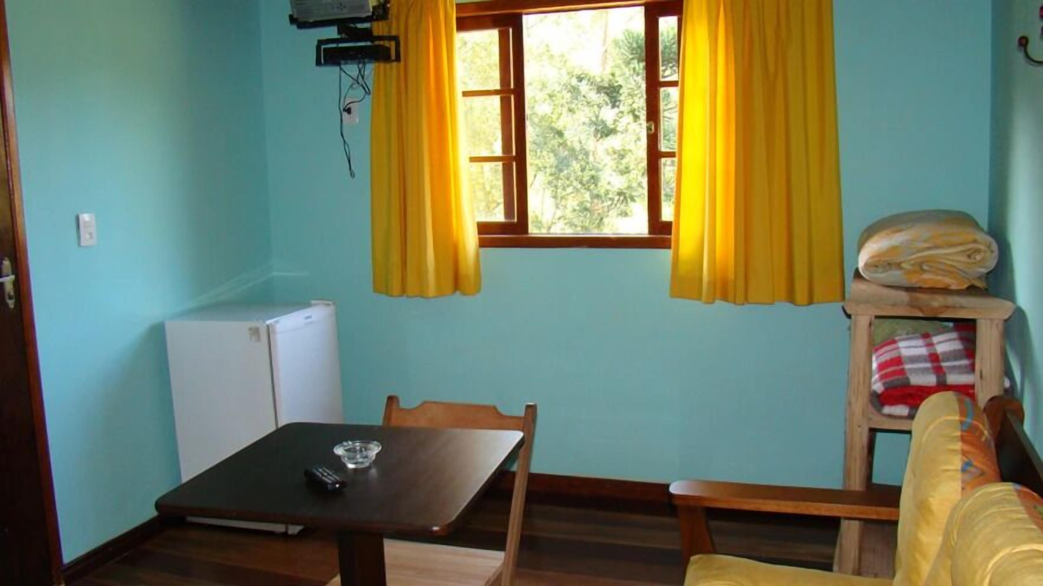 Una habitación pequeña con una mesa, sillas, un sofá, un ventanal con cortinas amarillas, un refrigerador blanco y un armario de madera con mantas dobladas.