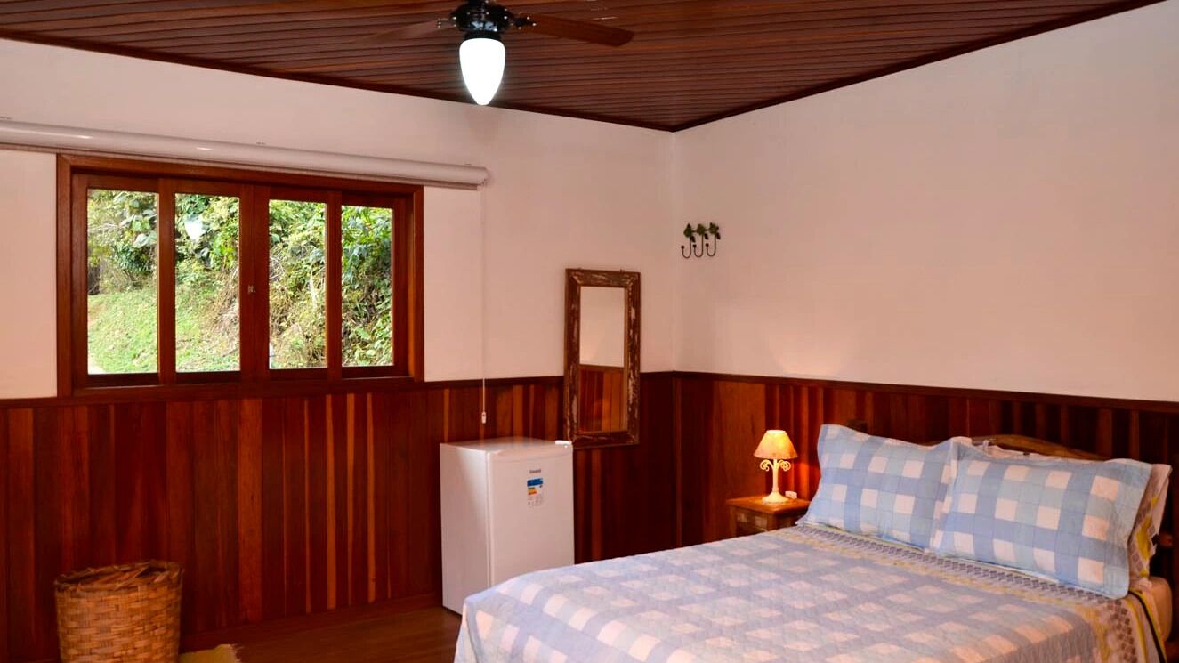 A bedroom with a bed, a small table with a lamp, a mirror, and a window with a view of greenery.