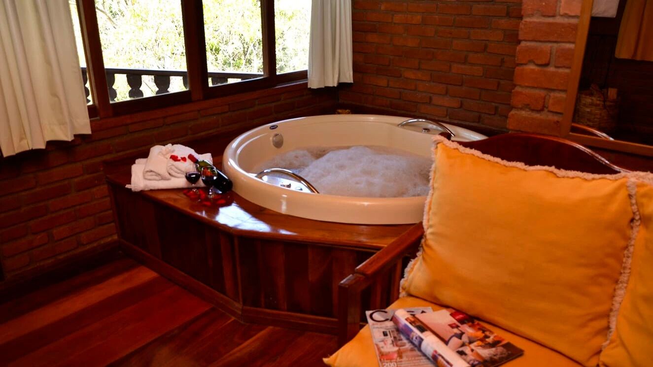 Una habitación con piso de madera, una bañera grande llena de burbujas, un banco de madera, un cojín amarillo y una mesa con una revista y una botella.