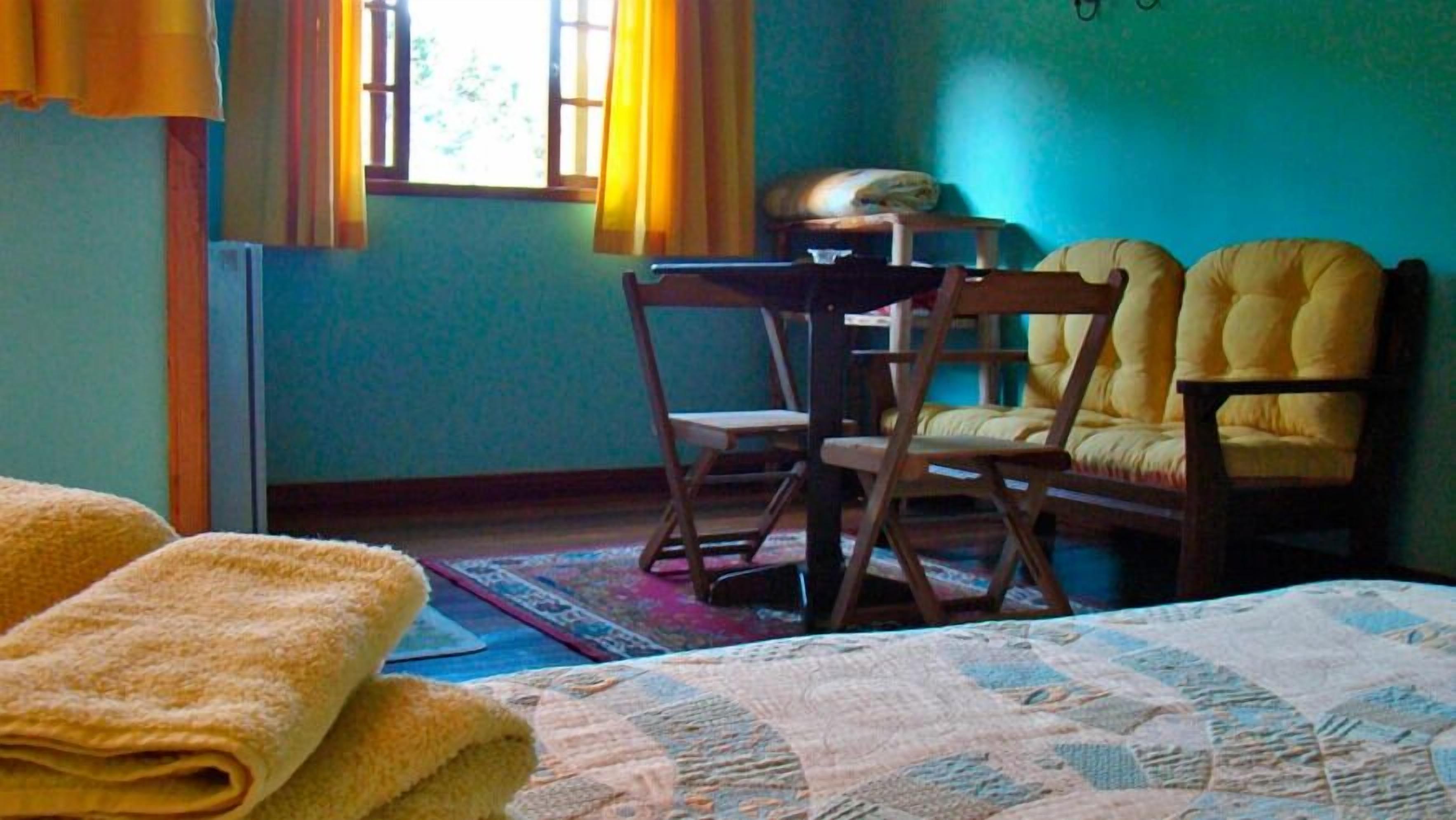 A room with a bed, two yellow armchairs, a wooden table, and a window with curtains.