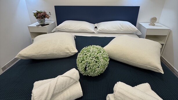 A neatly made bed with pillows and towels, a decorative flower arrangement, and a painting of the Colosseum on the wall.