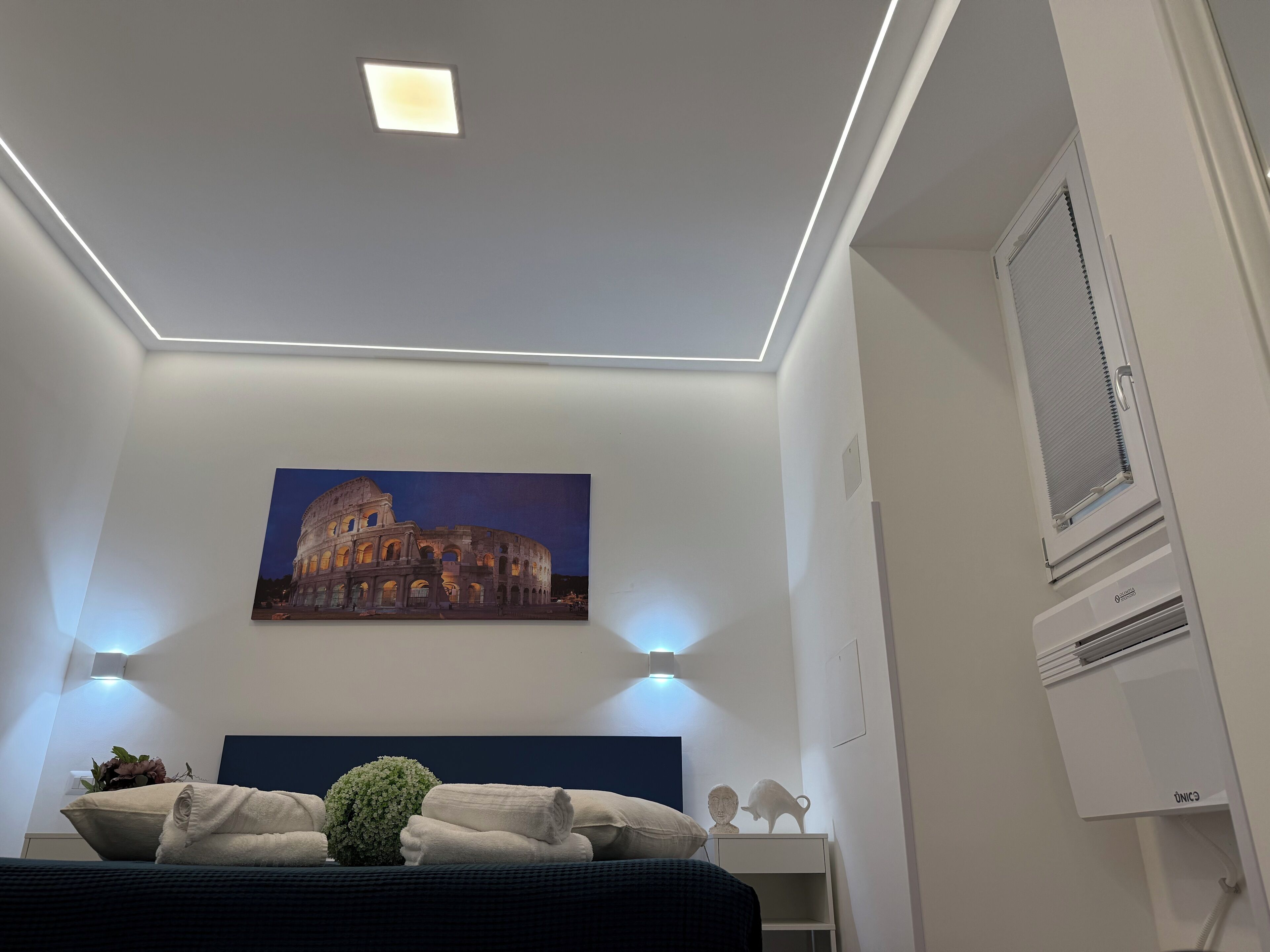 A bedroom with a bed, a painting of the Colosseum, a window with blinds, and a ceiling light.