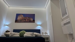 A bedroom with a bed, a painting of the Colosseum, a window with blinds, and a ceiling light.