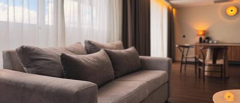 A modern living room with a grey sofa, a dining area with a table and chairs, and a large window with sheer curtains.