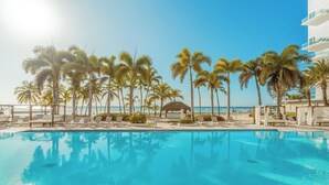 A swimming pool with lounge chairs and palm trees, a beach in the background, and a building with balconies.