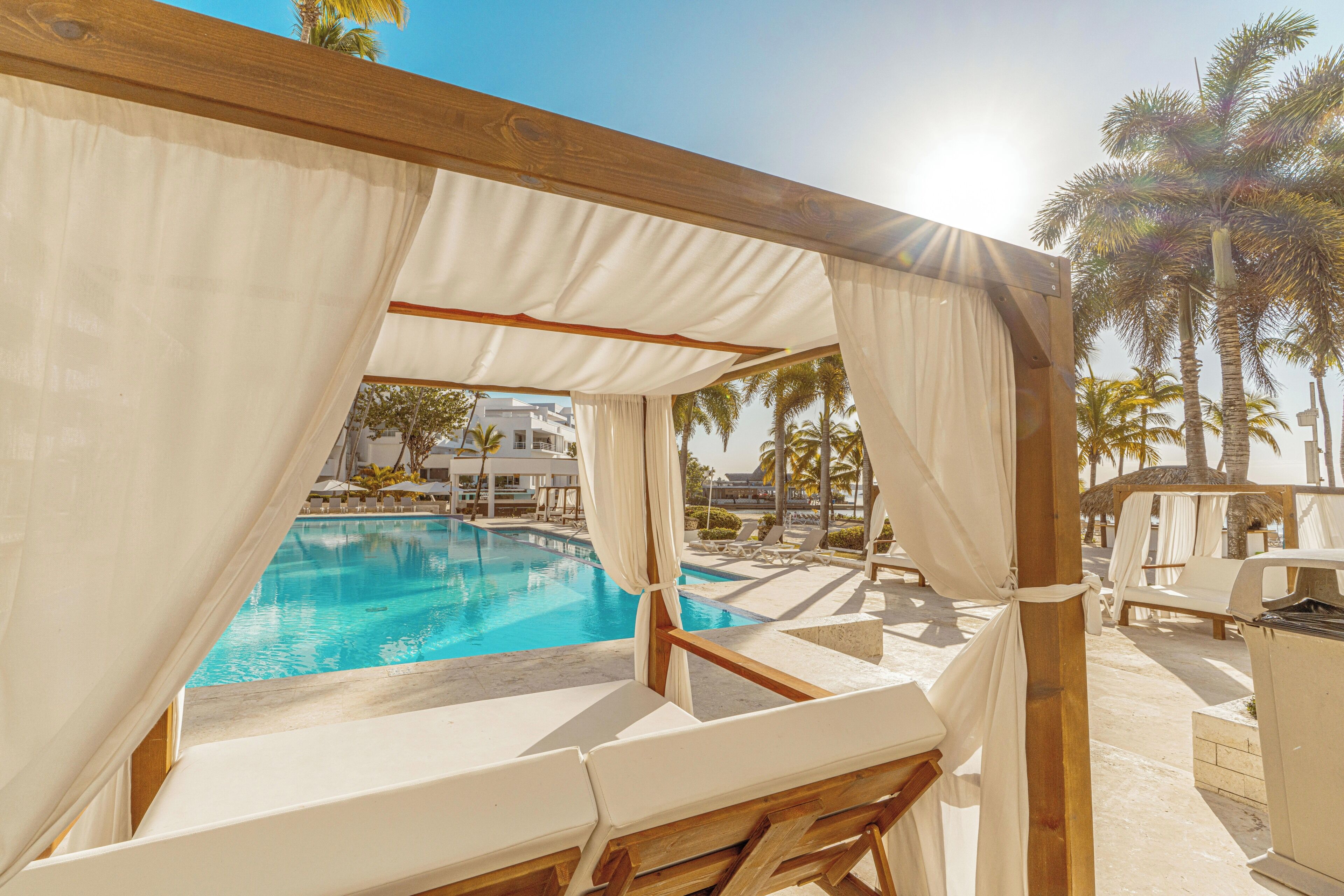 A poolside area with a covered seating area, a pool, and palm trees in the background.