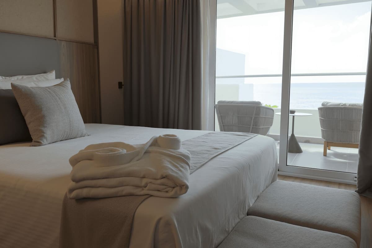 Quarto de hotel com uma cama grande, cabeceira, vista para o mar através de uma varanda e uma cadeira perto da janela.