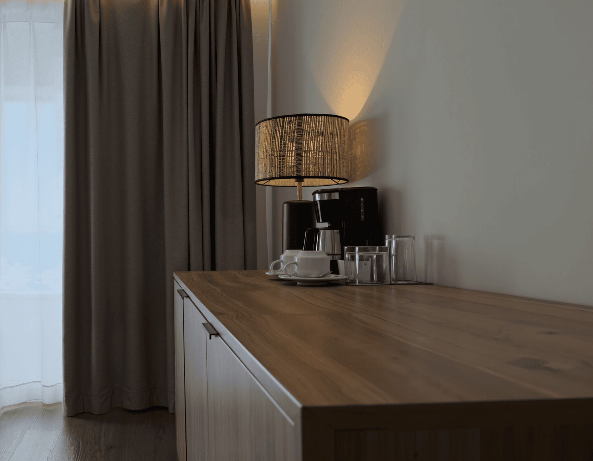 A wooden desk with a coffee maker, cups, and glasses, a lamp, and a window with curtains.