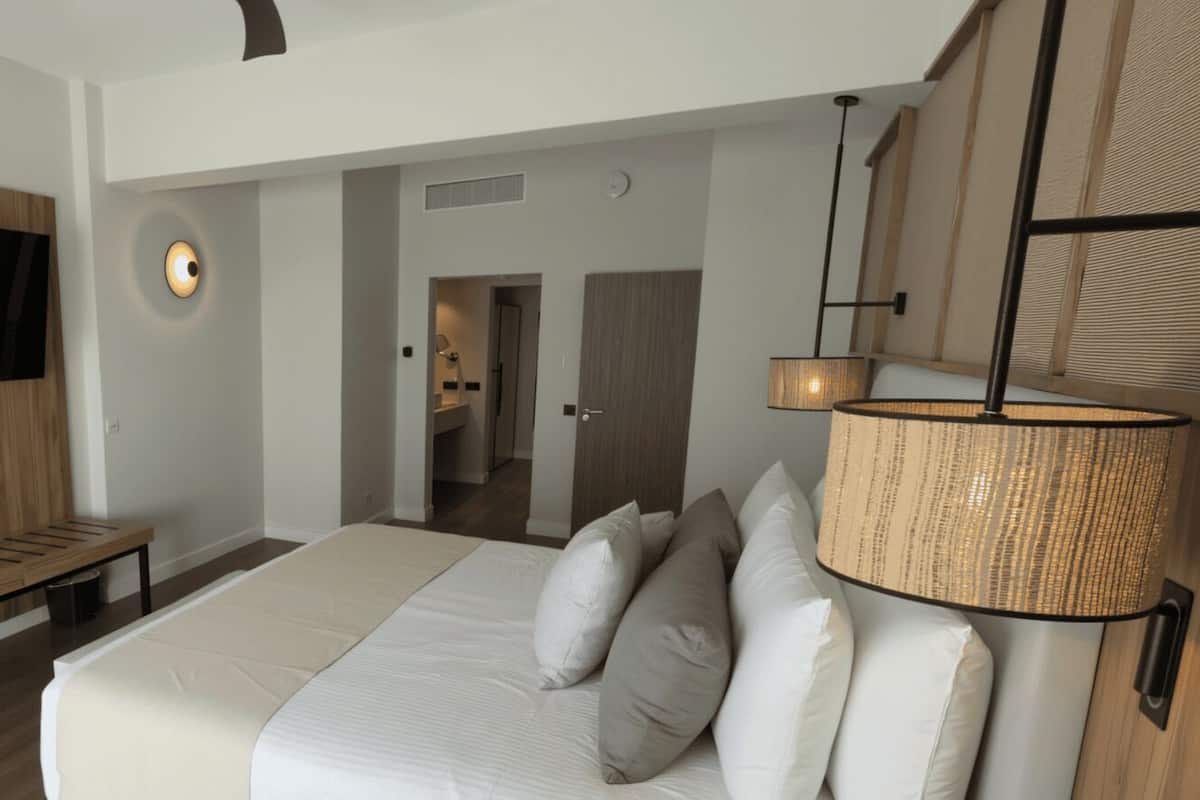 Quarto de hotel moderno com uma cama grande, um banco de madeira, uma TV de tela plana fixada em um painel de madeira, uma luminária de parede e uma porta para um banheiro.