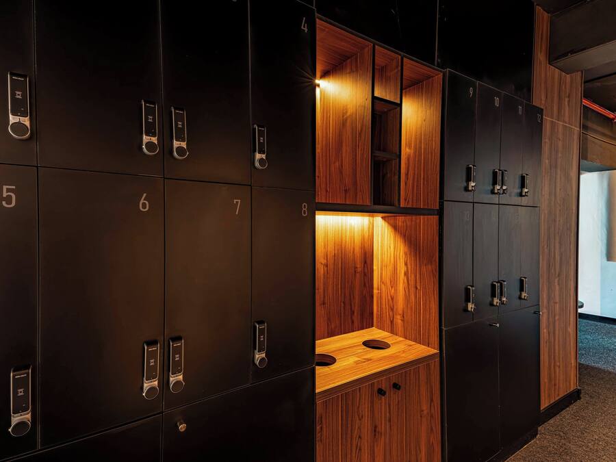 Een rij zwarte lockers met genummerde vakken, een houten wastafel met spiegel, en een blik op een gang.