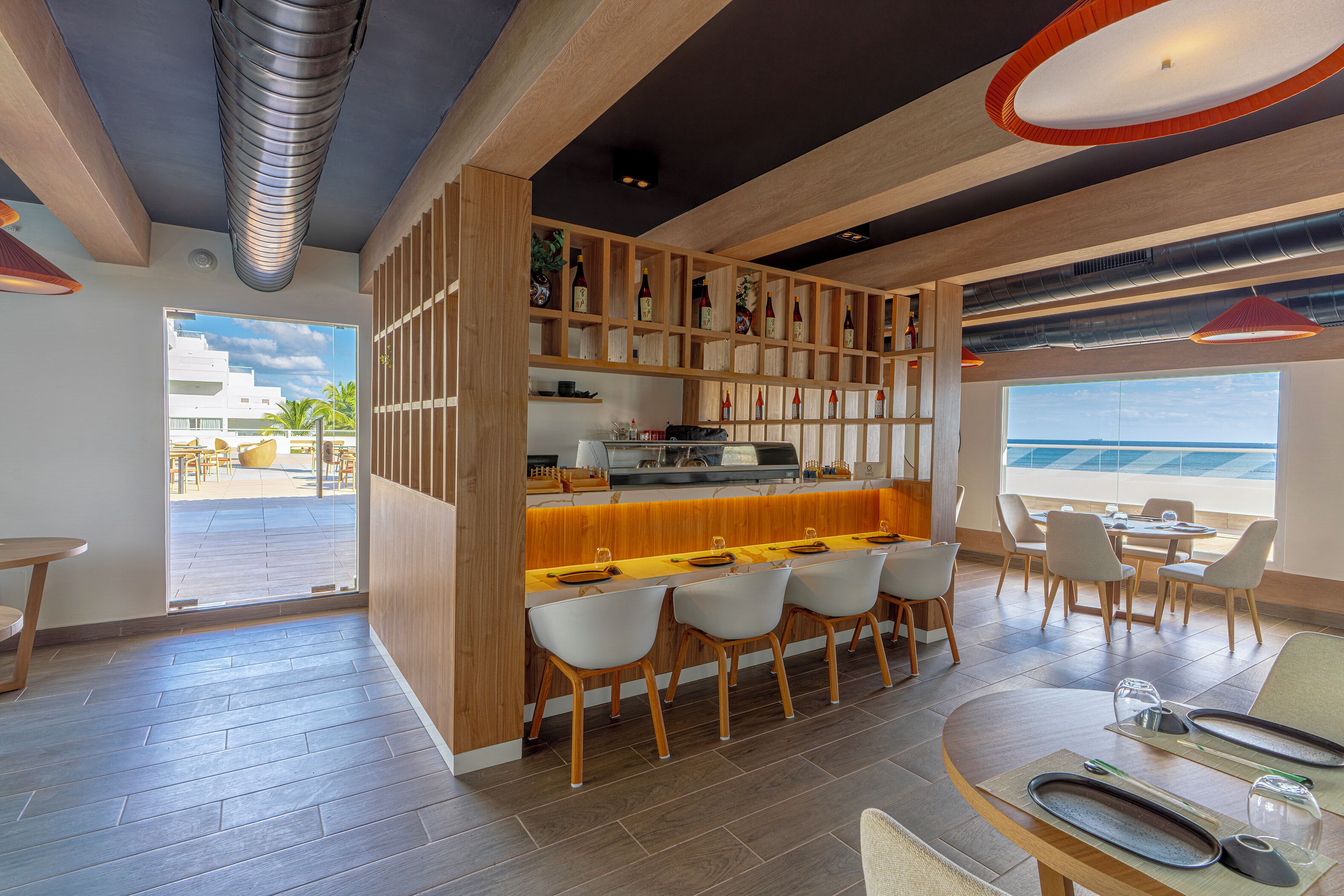 Un área de bar moderna con mostrador de madera, sillas blancas y vista al océano.