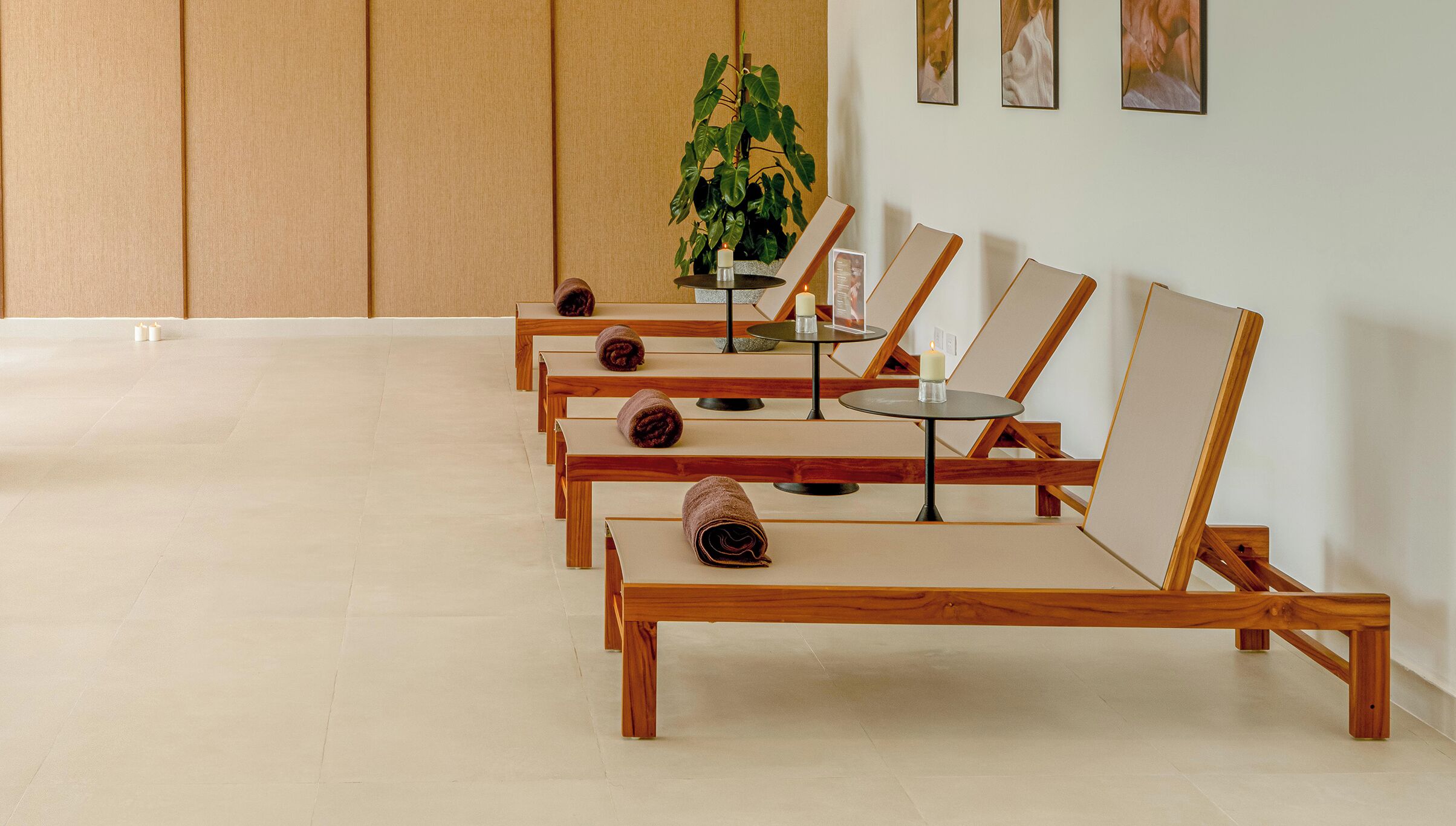A row of wooden lounge chairs with towels, a small table with a candle, and a potted plant in a room with a tiled floor and wooden panel walls.