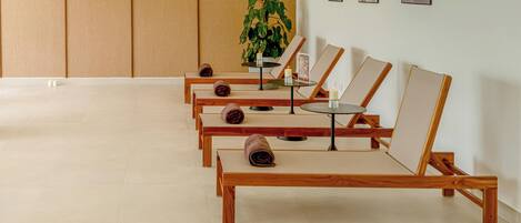 A row of wooden lounge chairs with towels, a small table with a candle, and a potted plant in a room with a tiled floor and wooden panel walls.