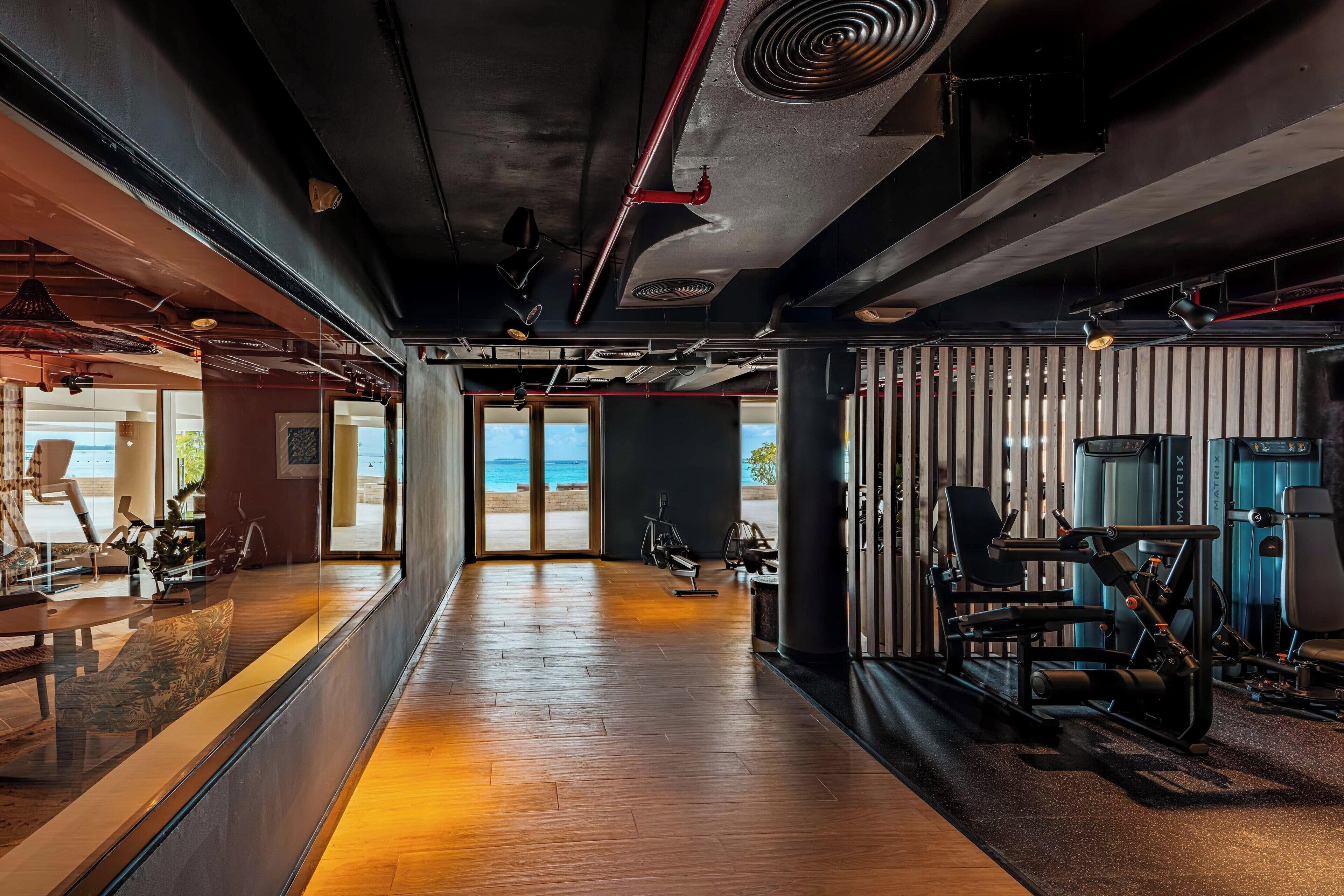 Een sportschool met trainingsapparatuur, een houten vloer en uitzicht op het strand door grote ramen.