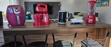 A modern kitchen with a marble countertop, a red coffee maker, a black electric kettle, a white food processor, and a red blender. There are three black bar stools in front of the counter.