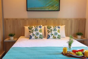Quarto de hotel com cama, duas almofadas estampadas com flores, mesa de cabeceira com planta, quadro na parede e bandeja de café da manhã com frutas e suco.