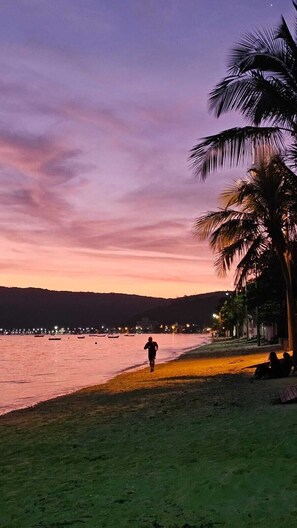 A beach scene with a person walking along the shore, palm trees, and boats in the water.