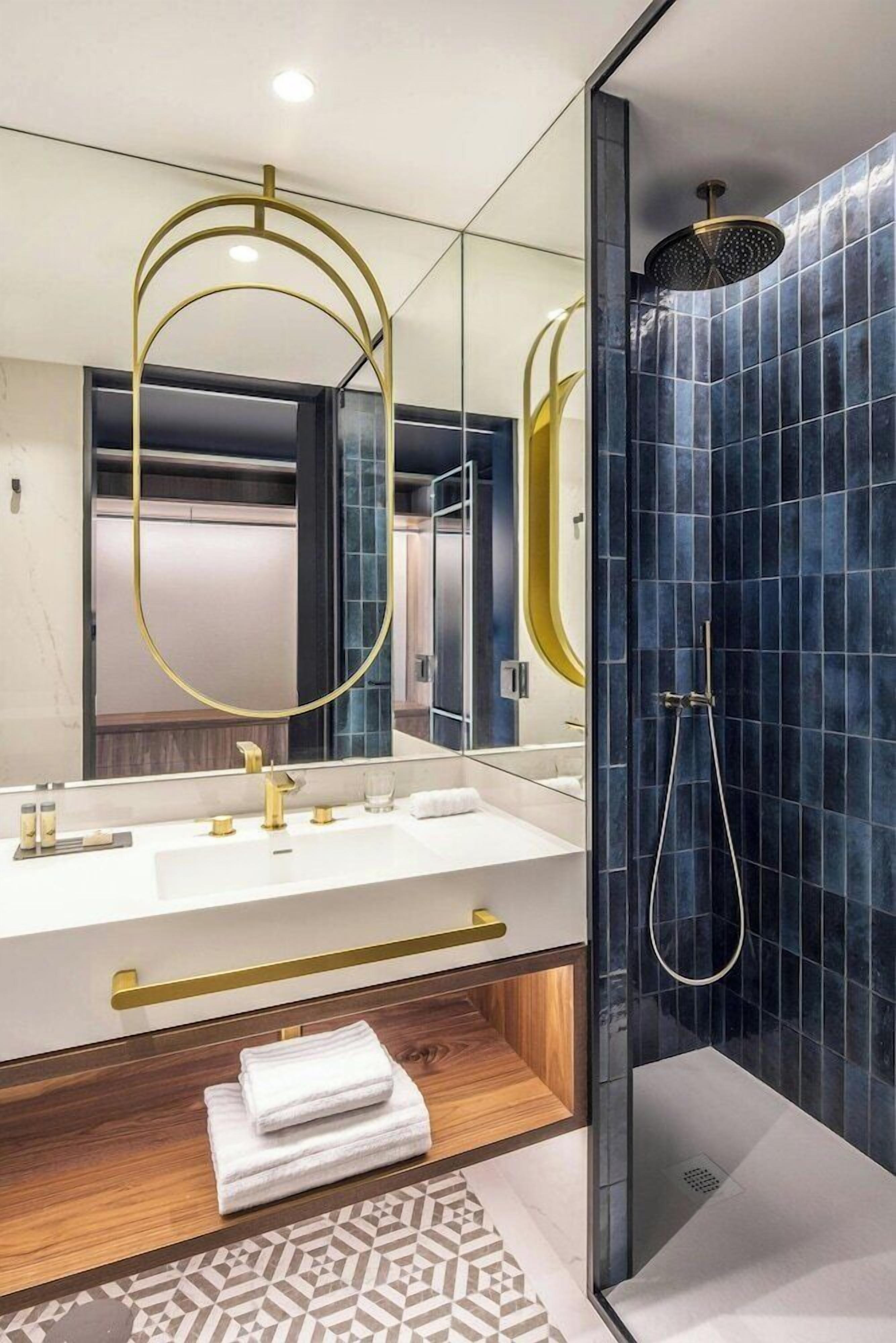A modern bathroom with a large mirror, two sinks with gold fixtures, a shower with a gold showerhead, and a patterned floor.