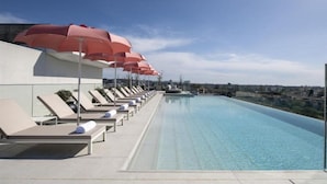 Una piscina a sfioro sul tetto con poltrone relax e ombrelloni rossi, con vista sulla città.