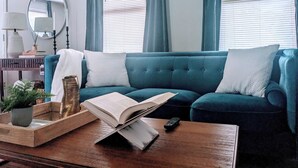 Ein blaugrünes Sofa mit weißen Kissen, ein Couchtisch aus Holz mit einem Buch und einer Fernbedienung, ein Spiegel und ein Blumentopf.