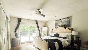 Ein Schlafzimmer mit Bett, Deckenventilator, Nachttisch, Lampe, Fenster mit Vorhängen und einer Tür, die auf einen Balkon führt.
