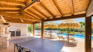 Ein Außenbereich mit Tischtennisplatte, Grill und Pool mit Sonnenschirmen.