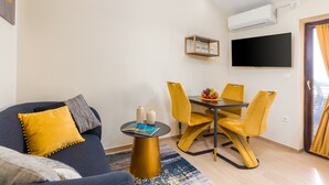 Ein modernes Wohnzimmer mit einer Couch, einem kleinen runden Tisch, einem Esstisch mit Stühlen, einem flachen Bildschirmfernseher an der Wand und einer Klimaanlage.