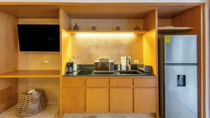 Cocina compacta con microondas, tostadora y refrigerador, armarios de madera y un televisor de pantalla plana montado sobre la encimera.