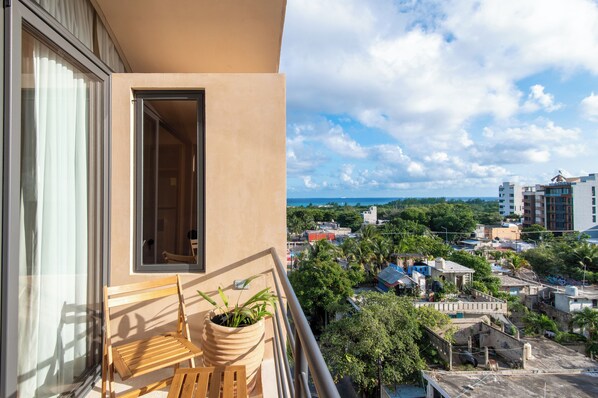 Un balcón con una silla de madera y una planta en maceta, con vistas a una zona residencial con varios edificios y vegetación.