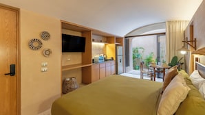 Un dormitorio con cama, televisión, kitchenette y balcón con plantas.