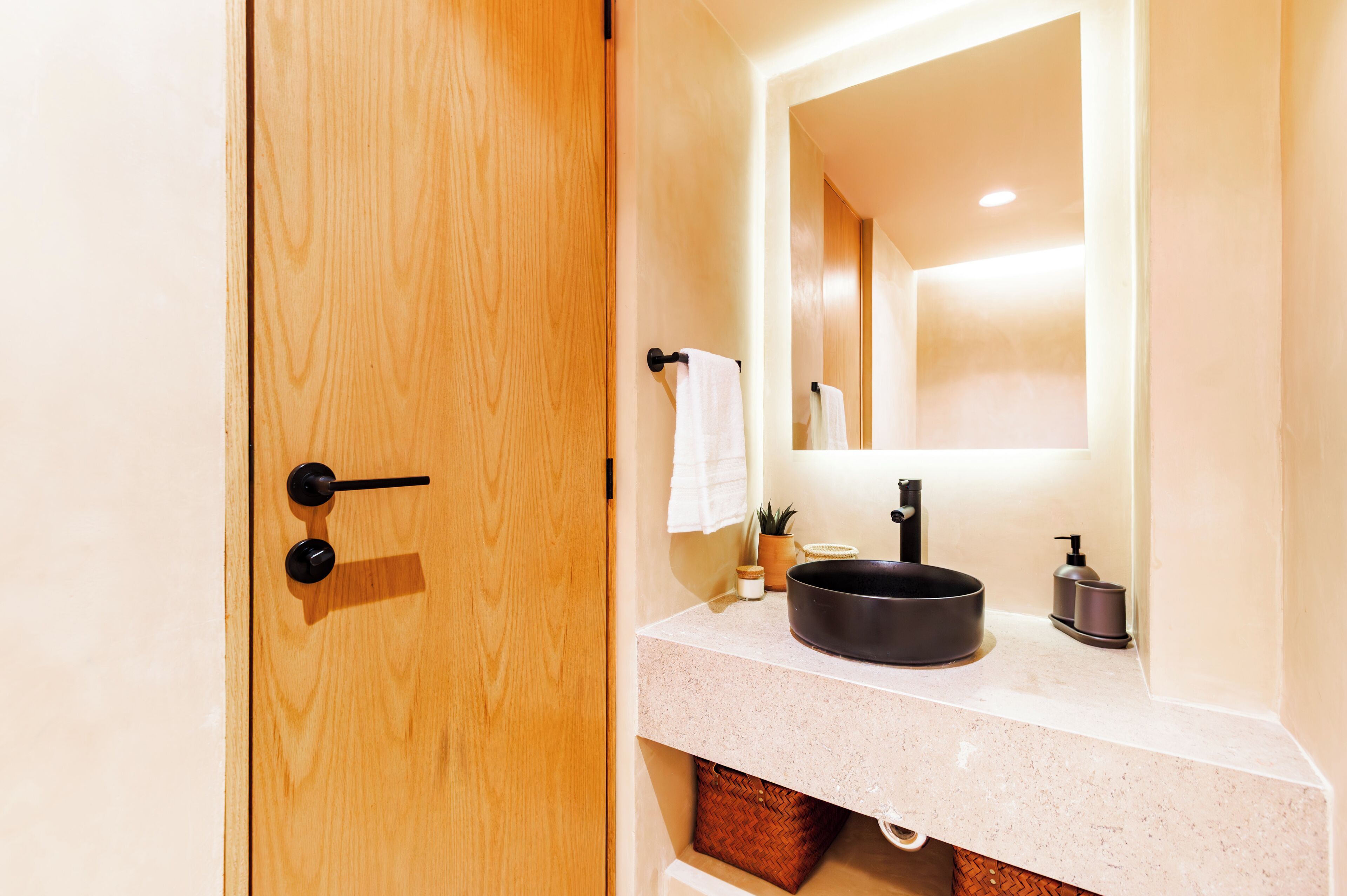 A bathroom with a wooden door, a sink with a black basin, a mirror, and a towel hanging on the wall.
