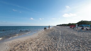 Una amplia playa de arena con cielo despejado, mar tranquilo y algunas personas.