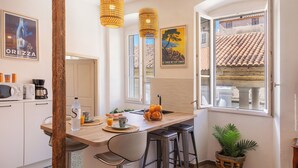 Una cocina con una mesa de madera, taburetes de bar, una cafetera, una botella y un bol con fruta.
