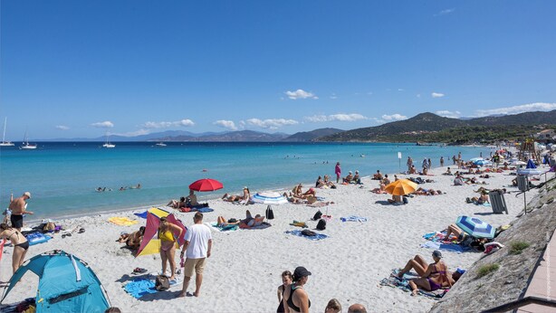 Una playa concurrida, gente tomando el sol y nadando, tiendas de campaña y sombrillas.