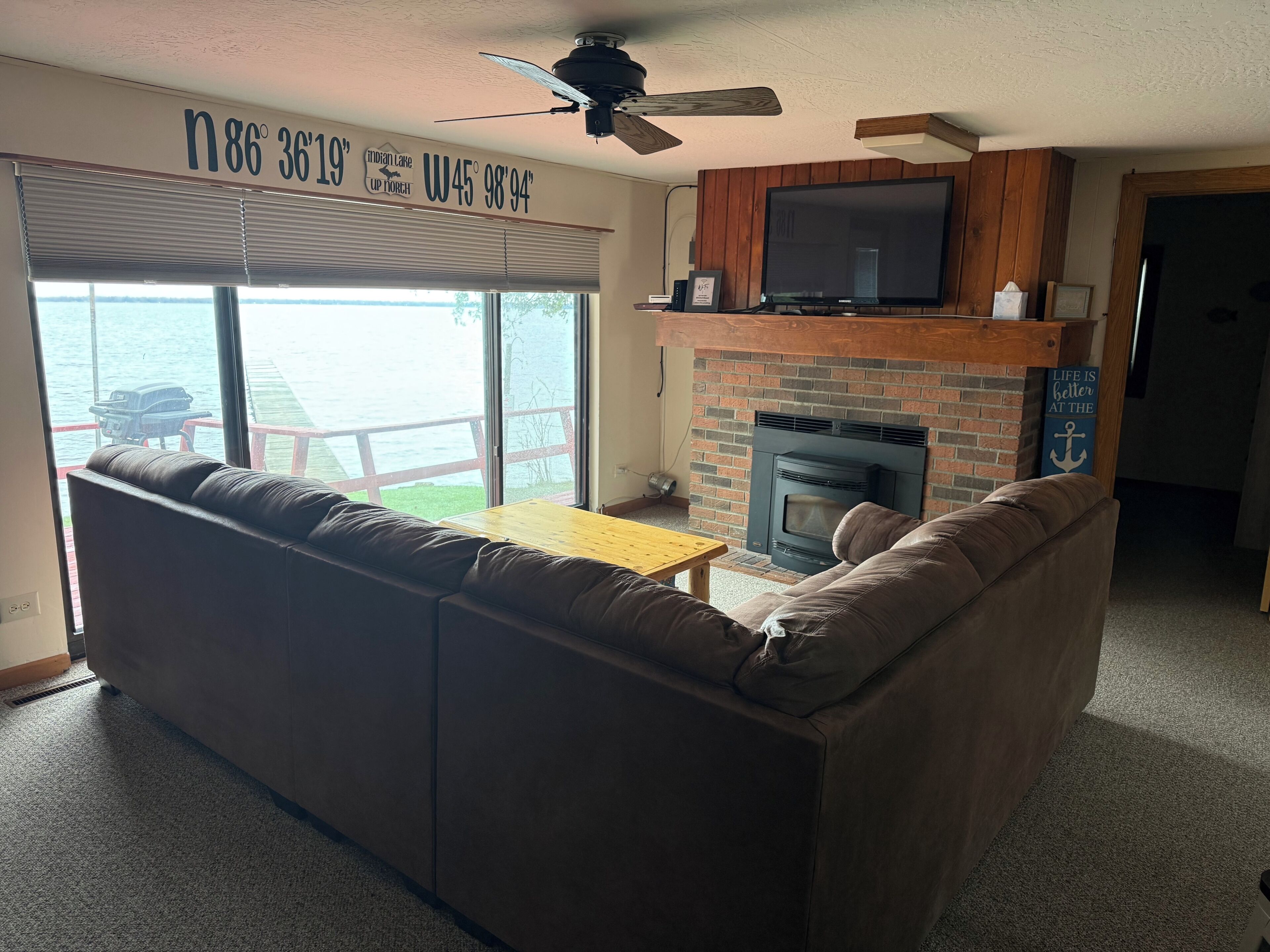 A living room with a large sectional sofa, a flat-screen TV mounted above a brick fireplace, and a ceiling fan.