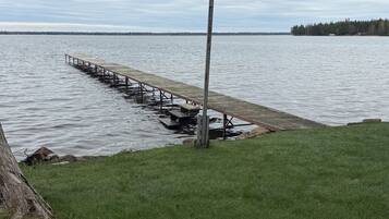 A wooden dock extending into a calm lake with a grassy shoreline and a tree in the foreground.